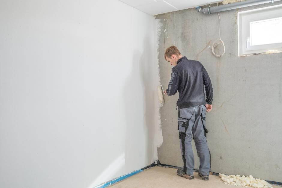 Junger Handwerker trägt mit Handrolle weiße Wandfarbe auf eine Kellerwand auf