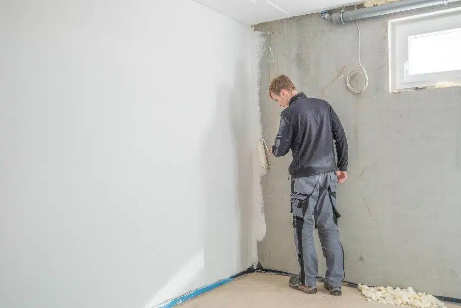 Junger Handwerker trägt mit Handrolle weiße Wandfarbe auf eine Kellerwand auf