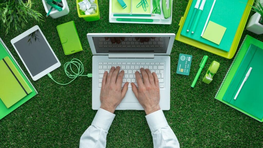 Blick von oben auf Laptop auf grünem Gras mit grünen Ordner, Schnellheftern und Büromaterialien um den Laptop herumverteilt.
