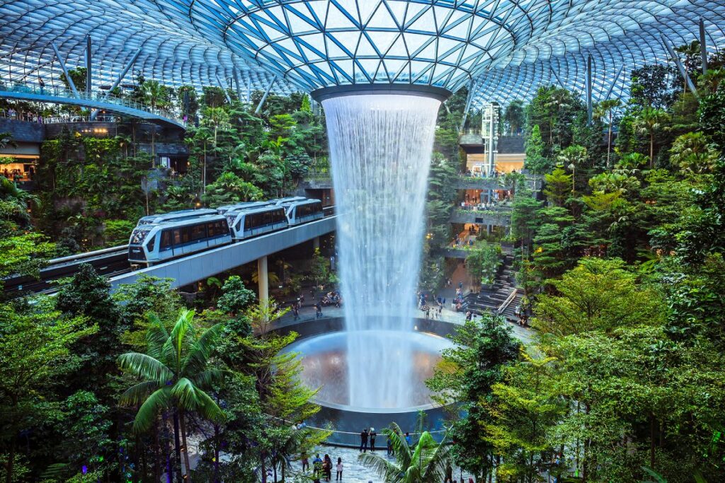 Trichterförmige Decke aus Glas. In der Mitte ist ein Indoor-Wasserfall, der von einem künstlichen Indoor-Dschungel umgeben ist. Menschen und ein Zug bewegen sich durch die Szenerie.